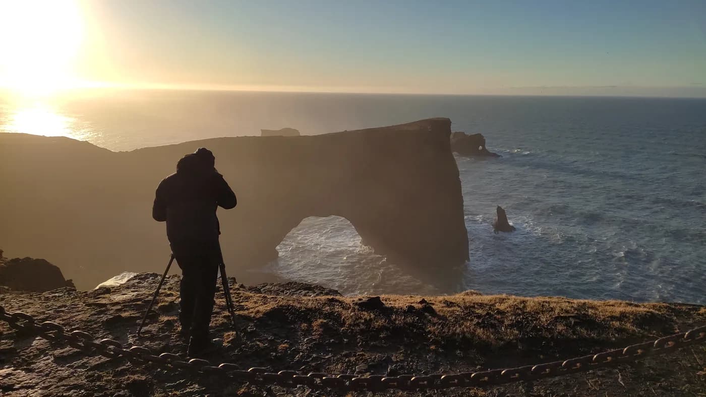 Fotógrafo con trípode en el acantilado de Dyrhólaey al amanecer con el arco natural de roca al fondo, sur de Islandia