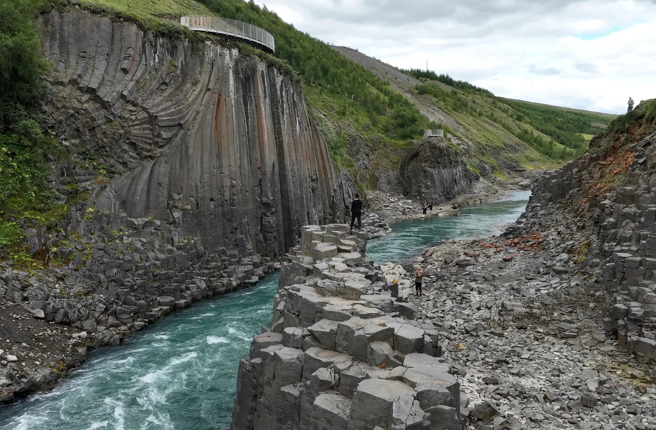 Composición fotográfica en el cañón de Stuðlagil, con personas dando escala junto al río y el mirador del lado oeste al fondo