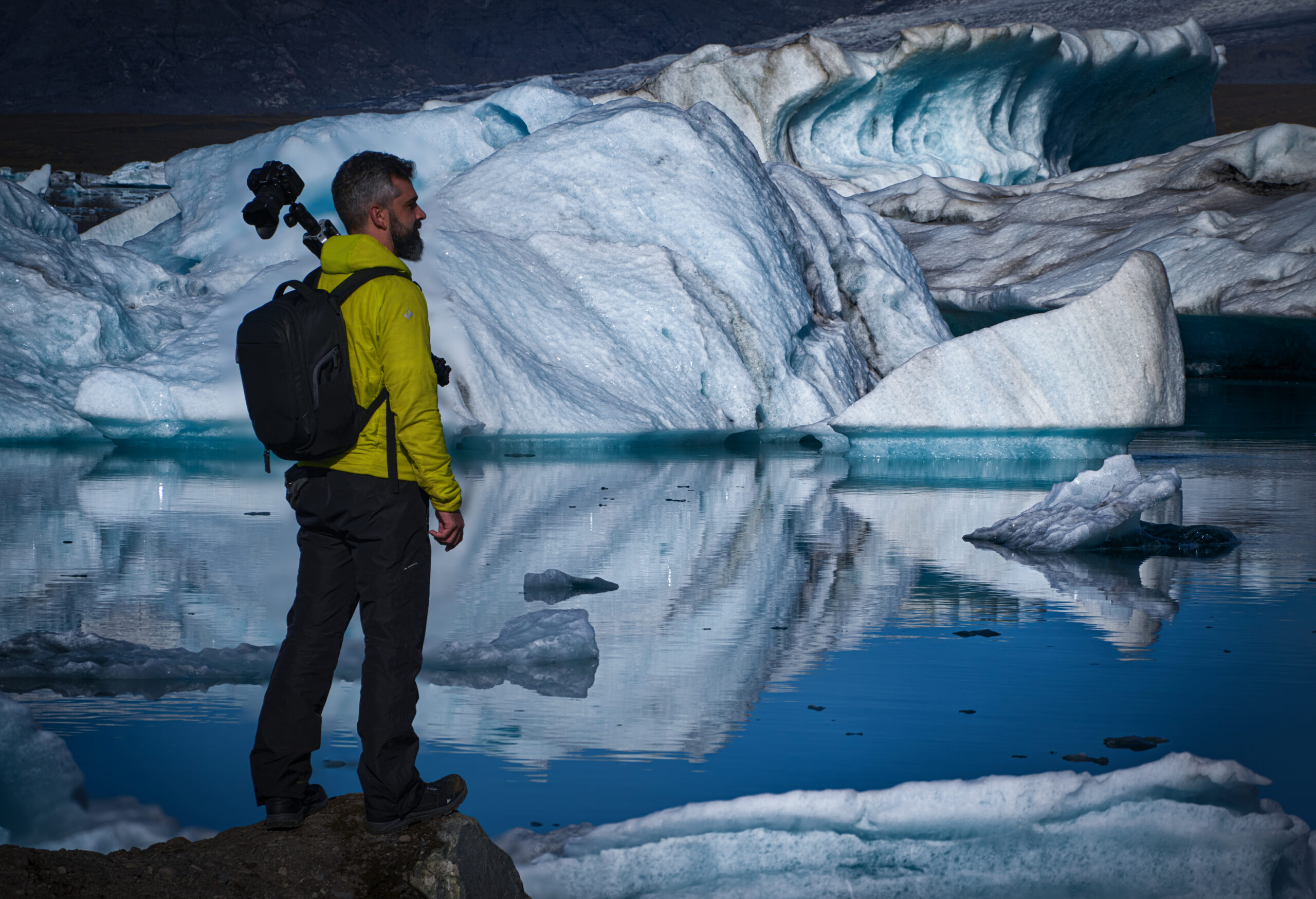 Fotógrafo con cámara y trípode al hombro frente a la laguna glaciar de Jökulsárlón con icebergs al fondo, Islandia