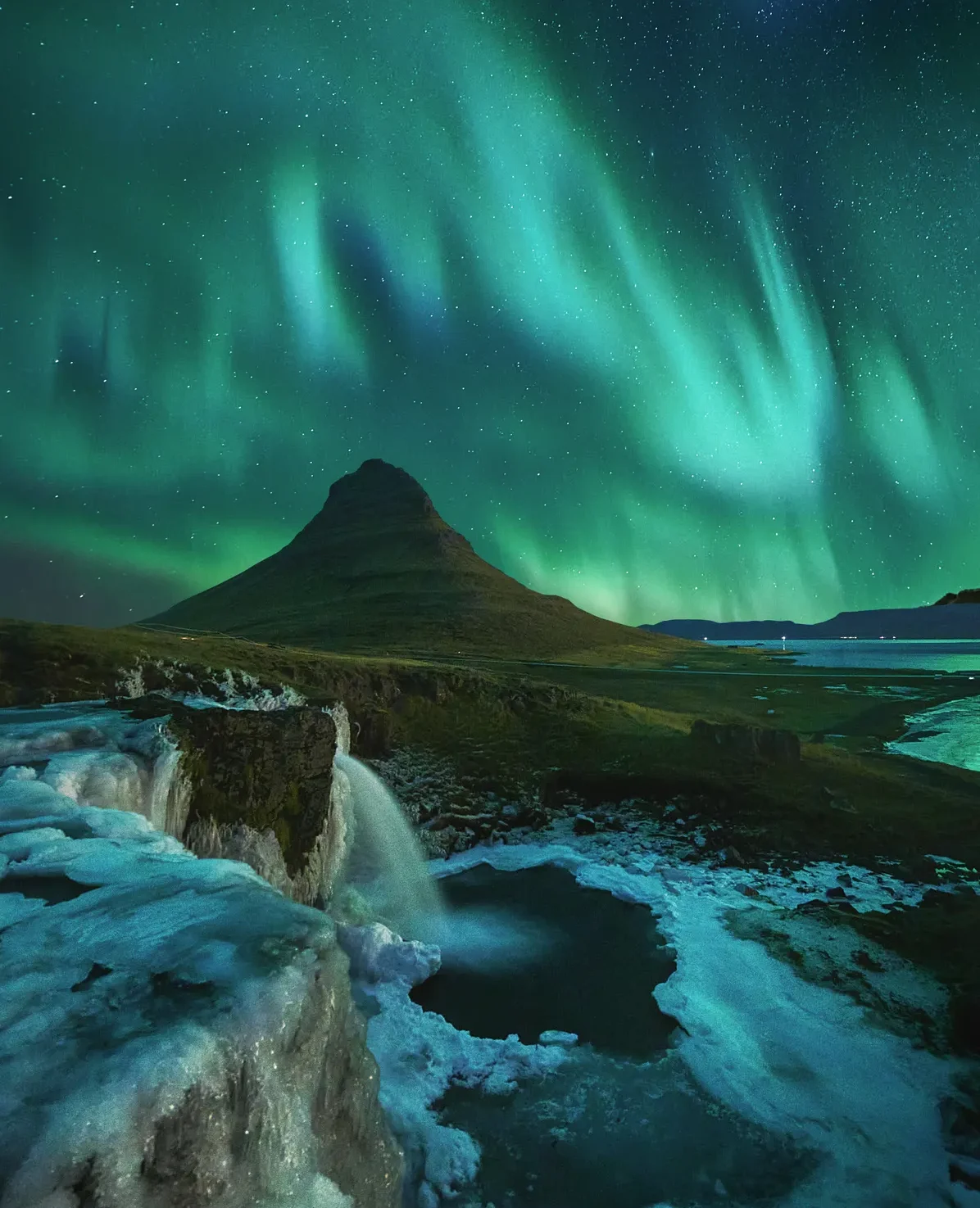 Kirkjufellsfoss helada con auroras boreales y Kirkjufell al fondo, Islandia Cascada Kirkjufellsfoss helada en larga exposición con el monte Kirkjufell al fondo bajo auroras boreales en snaefellsnes Islandia