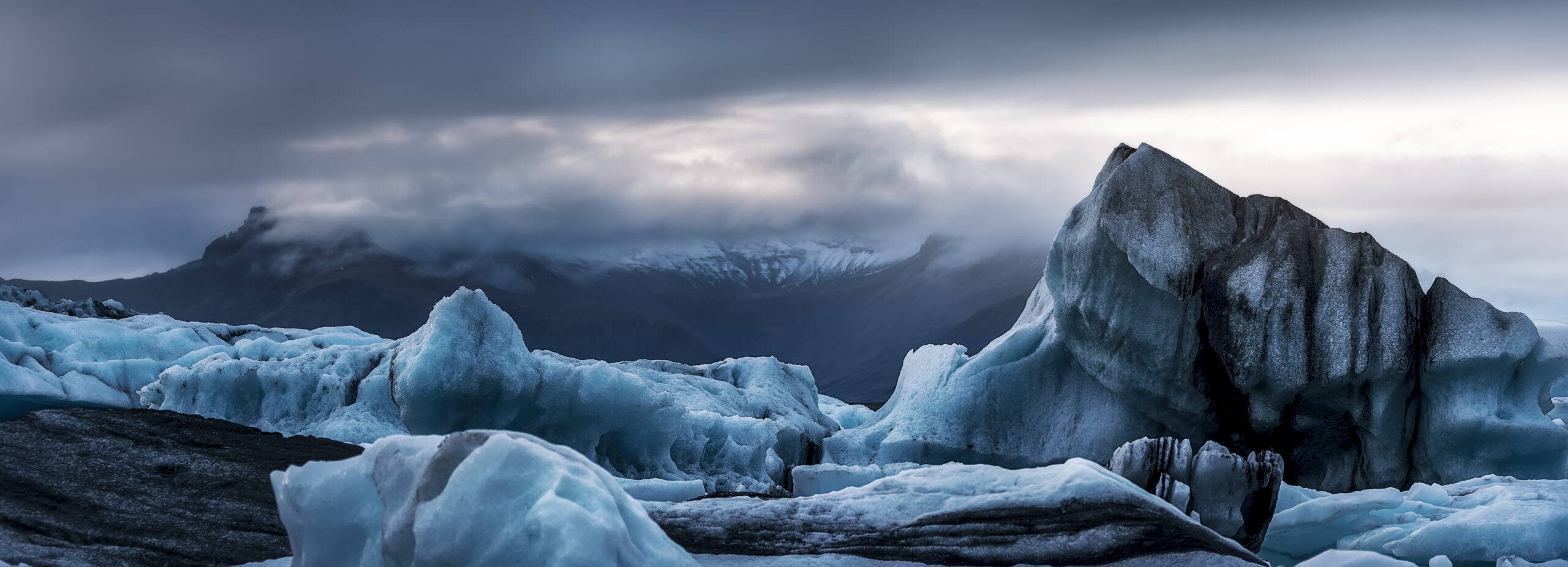 Panorámica de icebergs azules con vetas negras de ceniza volcánica en Jökulsárlón, montañas nevadas entre nubes al fondo, Islandia