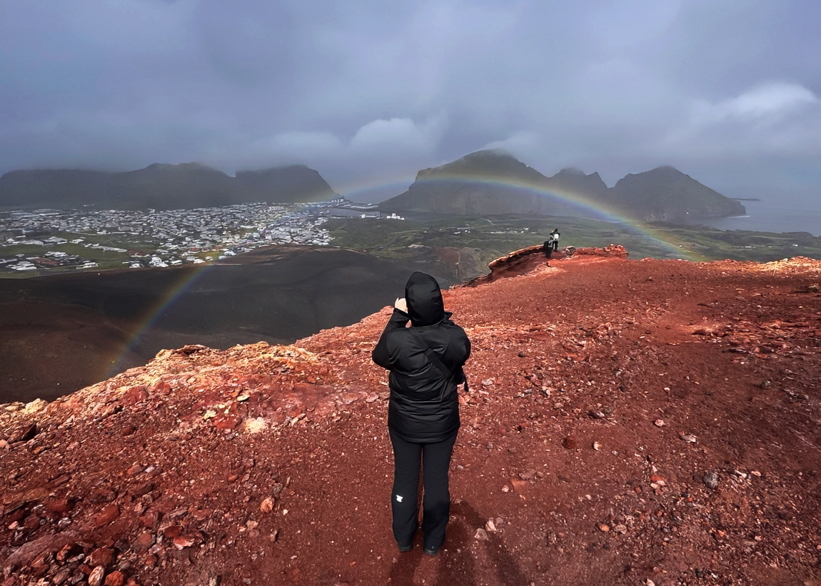 Persona fotografiando desde la cima del Eldfell con el pueblo de Heimaey y arcoíris al fondo